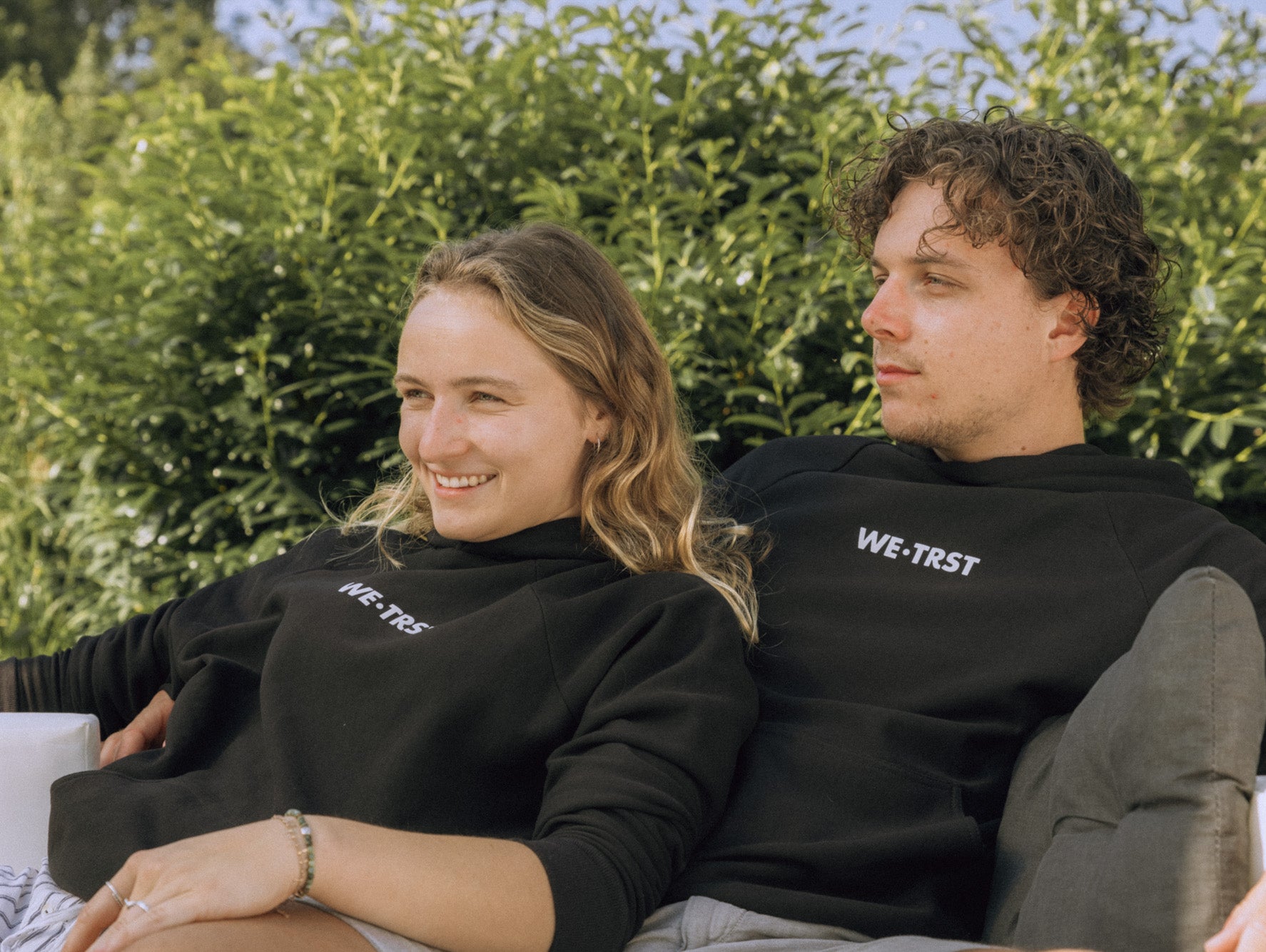 Das Bild zeigt ein Paar, das auf einem Sofa liegt und beide tragen einen schwarzen Hoodie von WE•TRST. Der Mann und die Frau liegen eng aneinander gekuschelt und scheinen eine entspannte Zeit miteinander zu verbringen.