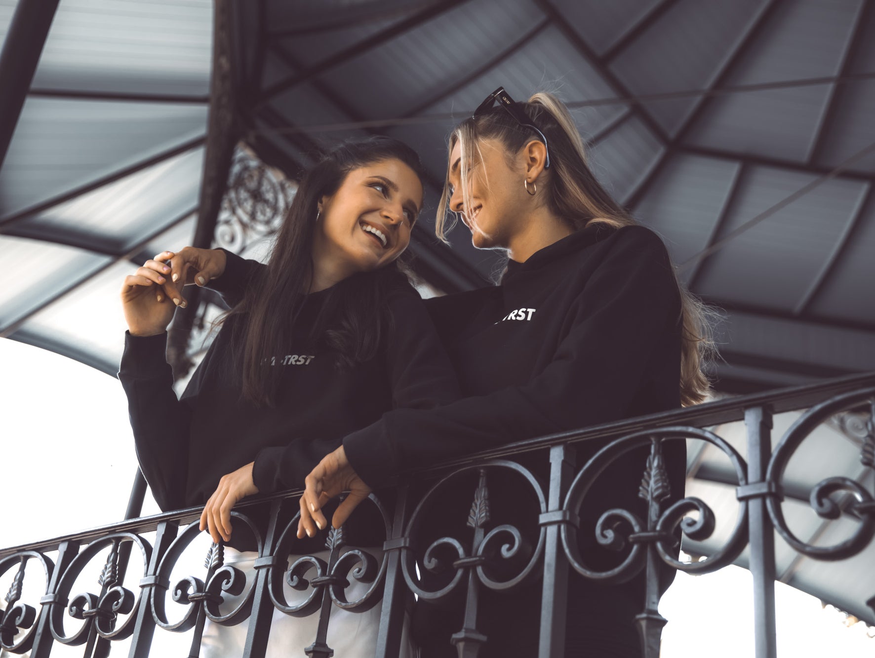 Das Bild zeigt zwei Freundinnen, die beide einen WE•TRST-Hoodie tragen und sich glücklich anschauen. Die Frauen stehen draussen in einer lebhaften Umgebung und haben ihre Arme um die Schultern des anderen gelegt.