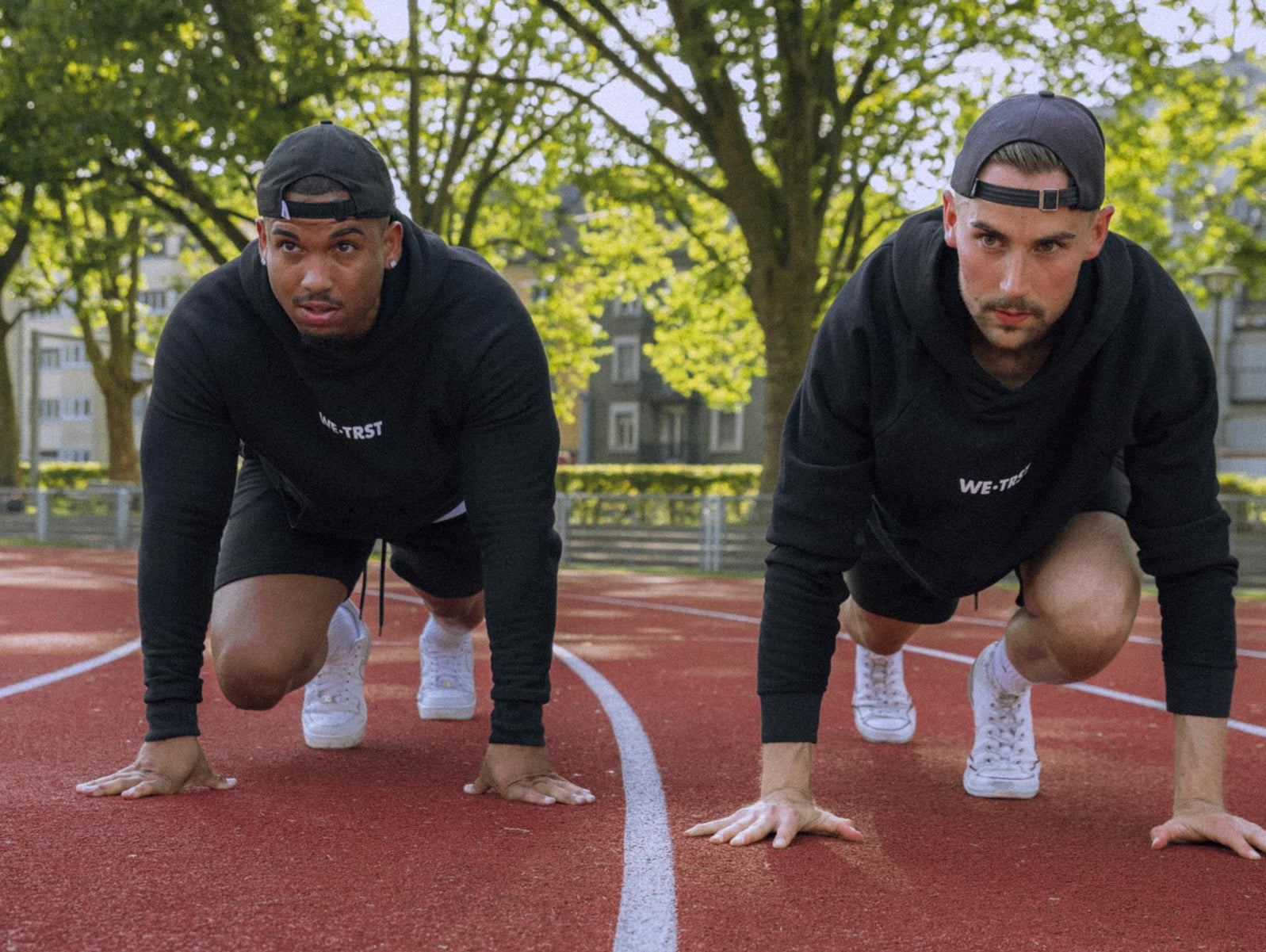 Das Bild zeigt zwei Sportler in Sprinter-Pose auf dem Boden, die beide schwarze Hoodies von WE•TRST tragen. Sie haben ihre Hände auf dem Boden platziert, während ihre Füsse in Startposition sind.
