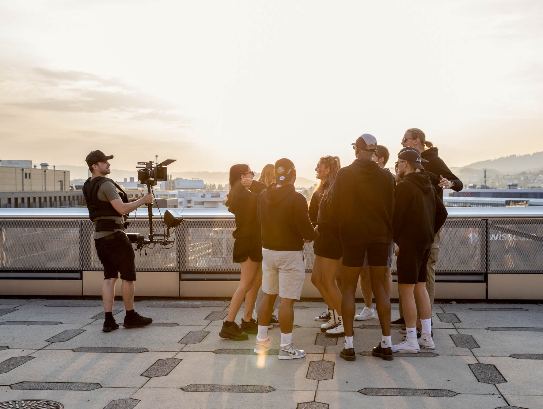 Eine Gruppe von Menschen hat auf einer Terrasse zusammen eine gute Zeit. Alle tragen schwarze Hoodies von WE•TRST. Sie lachen und unterhalten sich. Im Hintergrund ist die Sonne untergegangen und der Himmel ist in warmen Tönen von Orange und Rosa getaucht.