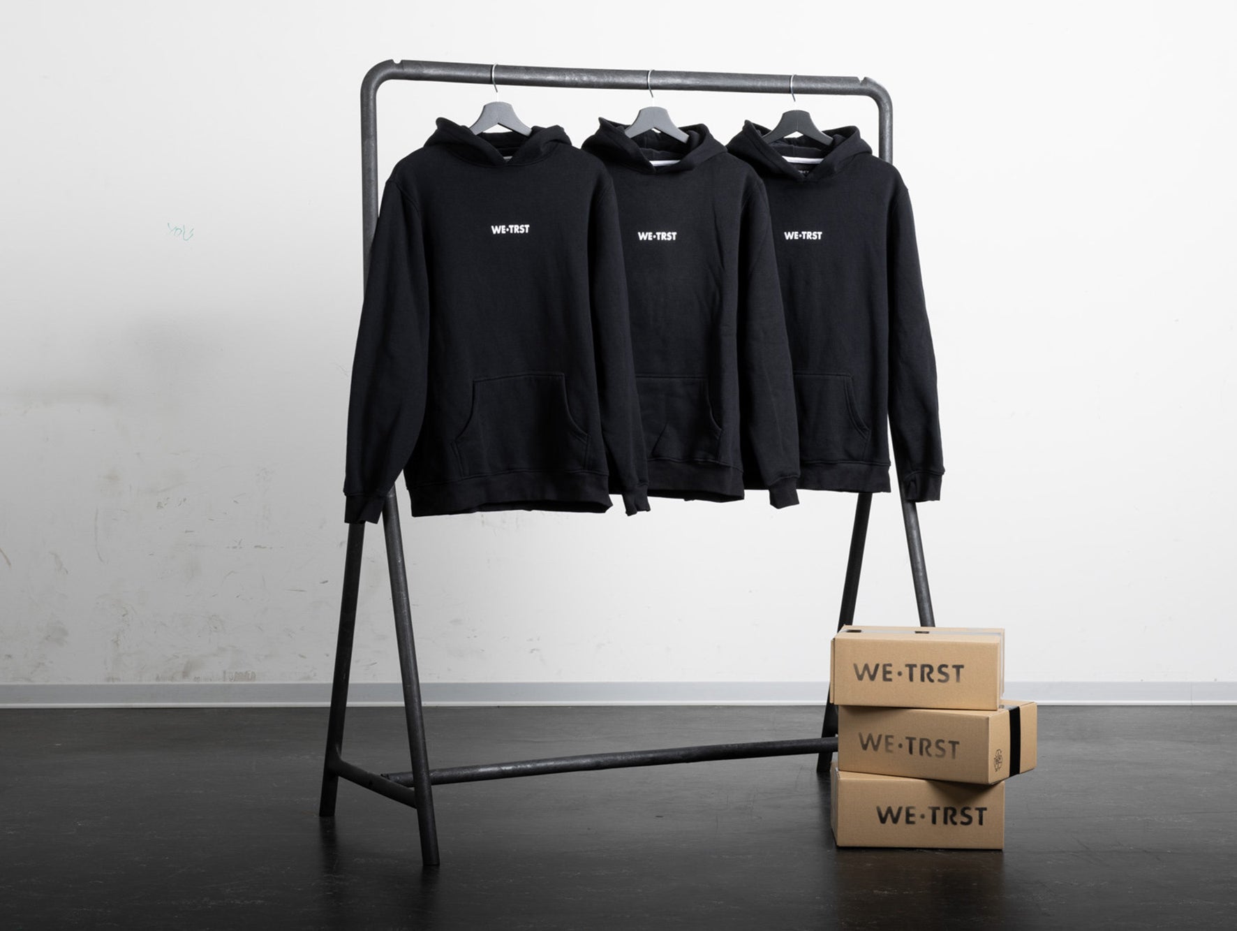 Drei schwarze Hoodies von WE•TRST hängen an einem Kleiderständer. Die Hoodies haben ein weisses WE•TRST-Logo auf der Brust.