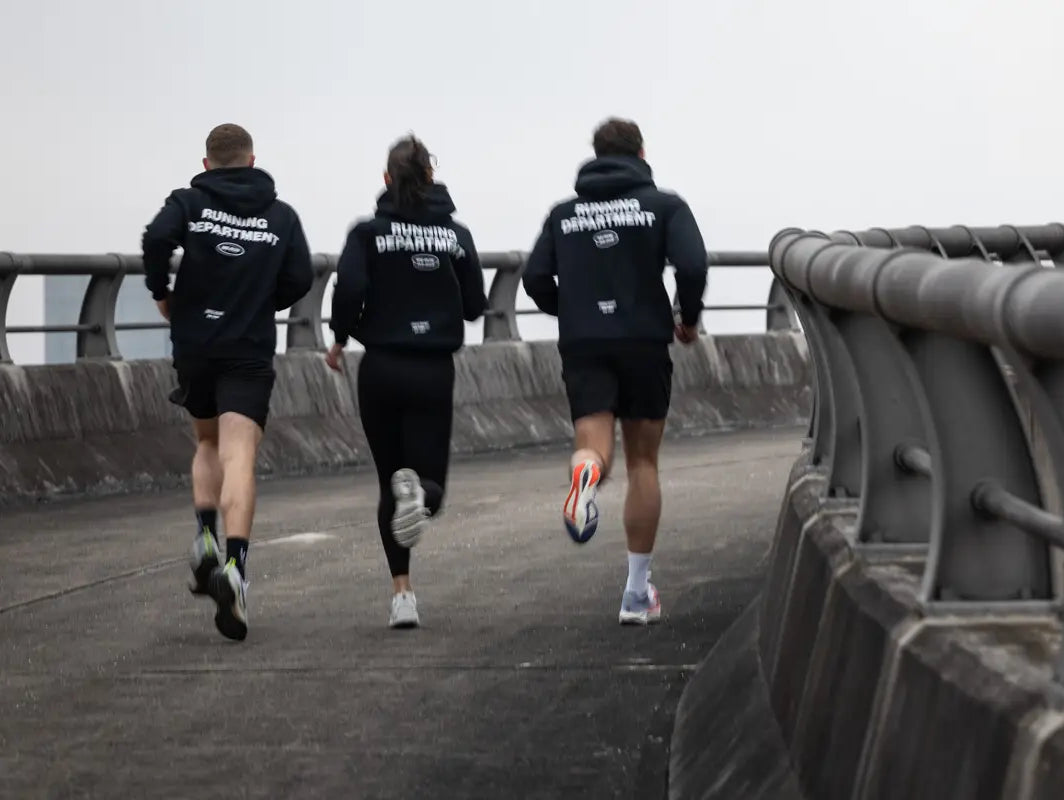 Drei Läufer:innen von WE•TRST laufen gemeinsam über eine Brücke, tragen schwarze Hoodies mit «Running Department» Backprint – Community Run, Urban Running, Laufcommunity Schweiz.