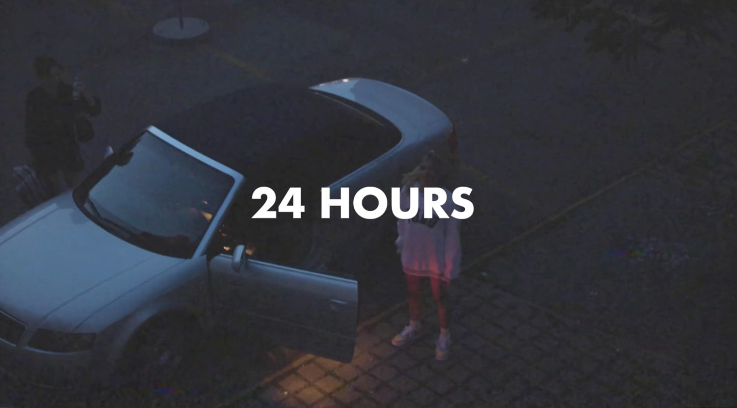 Das Bild zeigt ein Auto, das auf einem leeren Parkplatz geparkt ist. Neben dem Auto stehen zwei Personen und der Hintergrund ist unscharf. Über dem Bild steht der Titel "24 Hours", der auf das Thema des Videos hinweist.