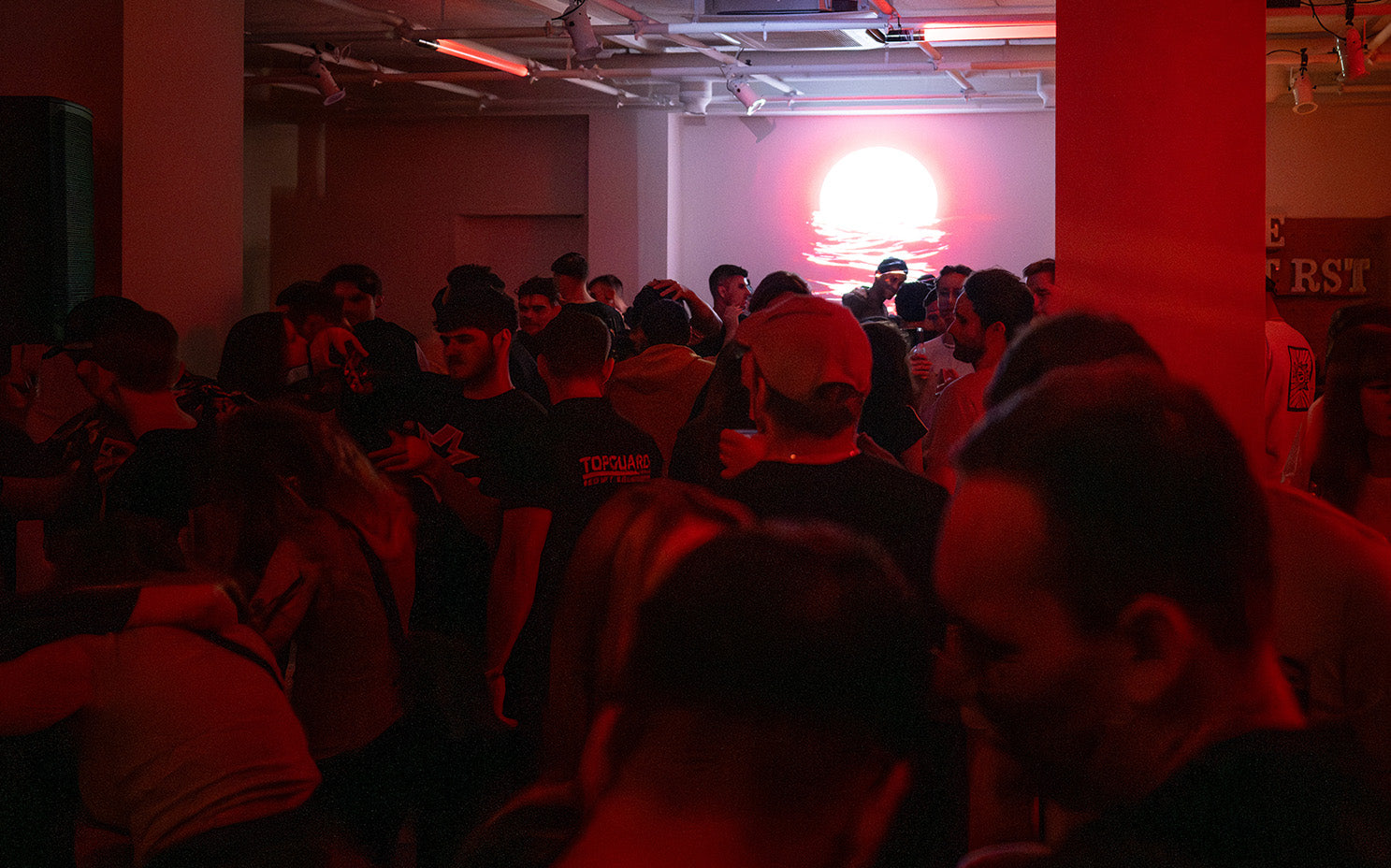 A crowded indoor rave of WE•TRST with red neon lighting and a sunset projection, showcasing the immersive experience of NOIR.