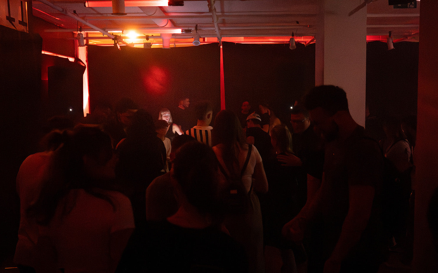 Tänzerinnen und Tänzer in einer schummrigen, in Rot- und Orangetöne getauchten Underground-Veranstaltung, die die NOIR-Rave-Atmosphäre von WE•TRST aufgreift.