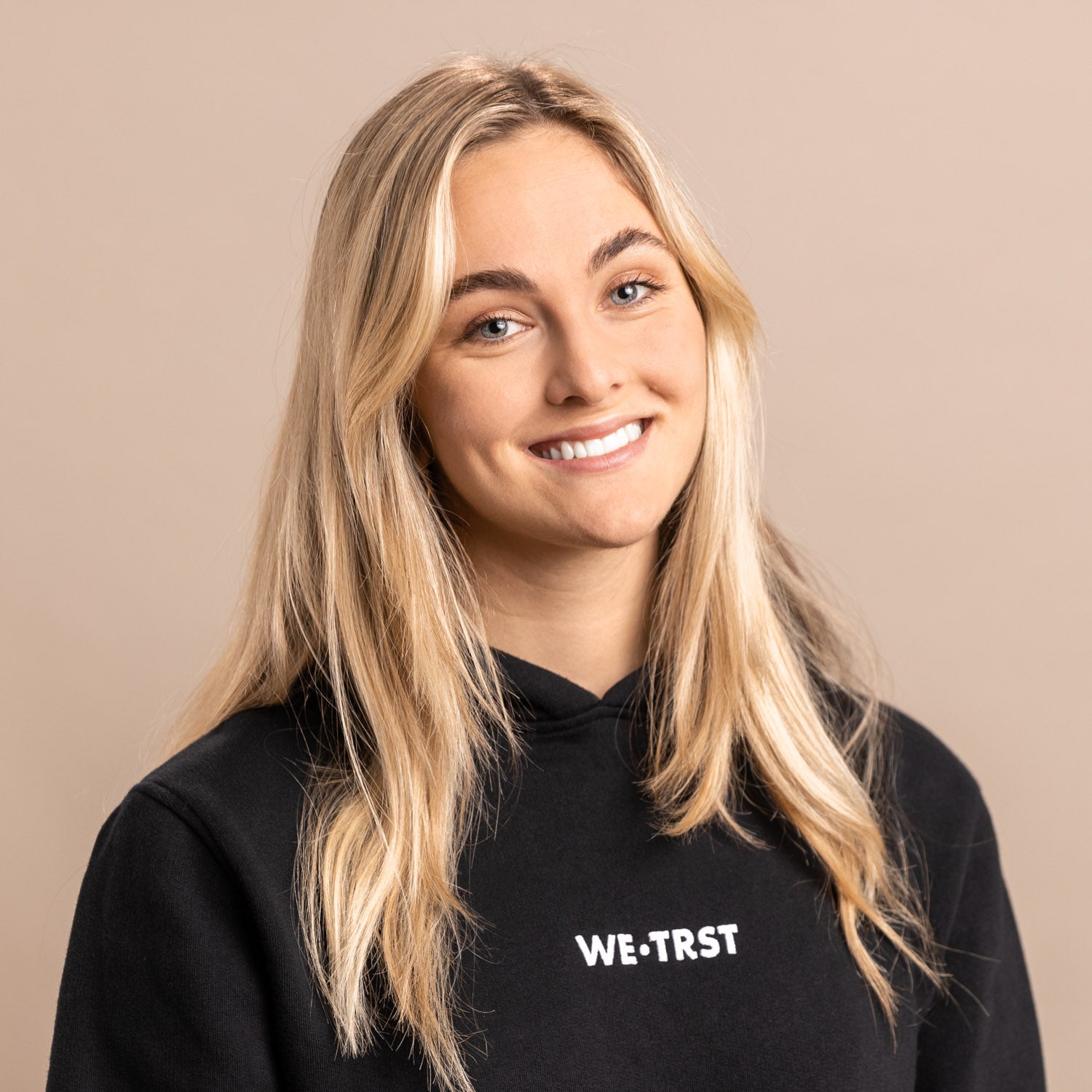 Das Bild zeigt Arina Luisa Bertényi, die Mitgründerin von WE•TRST. Sie steht im Studio und blickt direkt in die Kamera. Sie trägt einen schwarzen Hoodie der Marke, der mit dem markanten weißen Logo auf der Brust verziert ist.