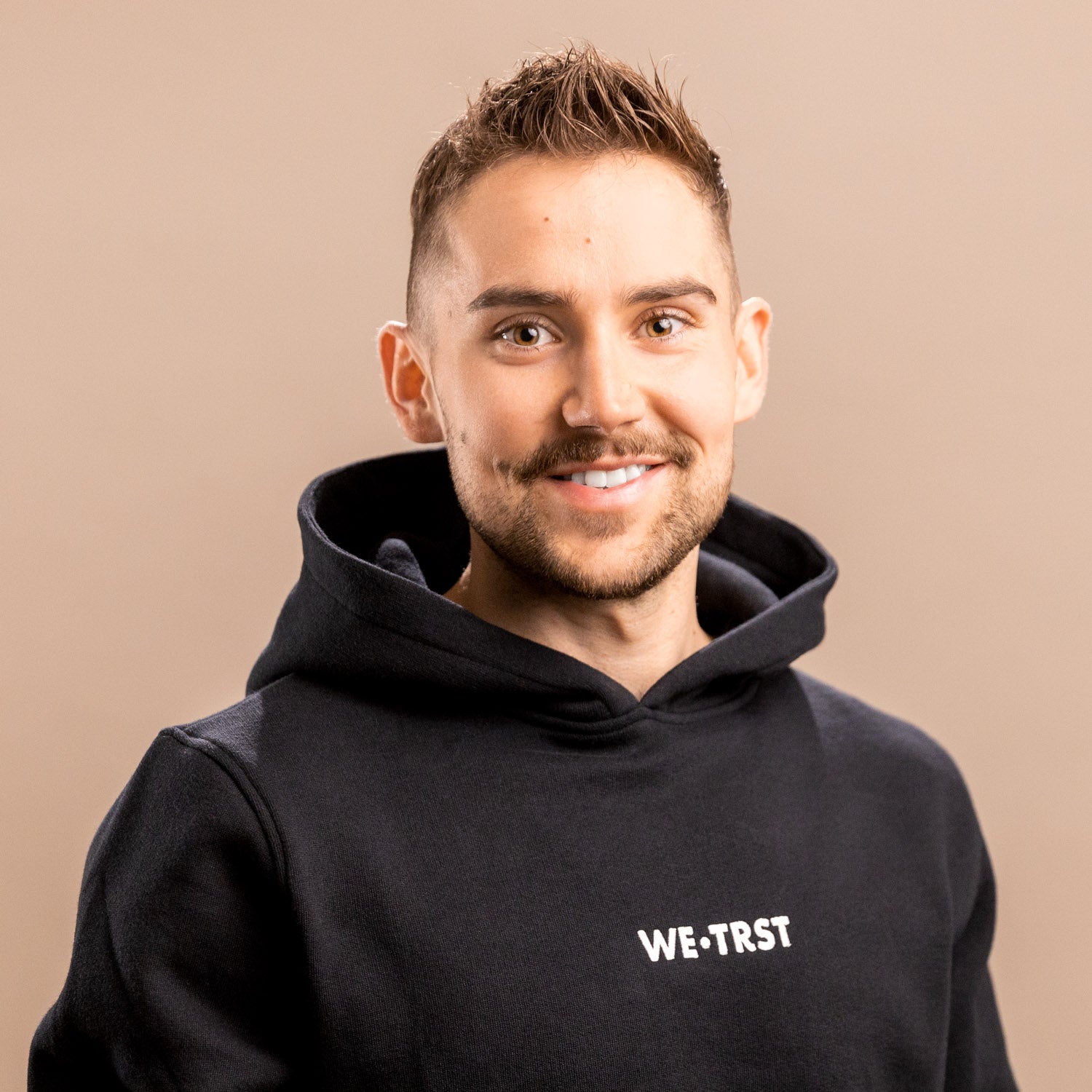 Das Bild zeigt Michael Müller, den Mitgründer von WE•TRST. Er steht im Studio und blickt direkt in die Kamera. Er trägt einen schwarzen Hoodie der Marke, der mit dem markanten weißen Logo auf der Brust verziert ist.