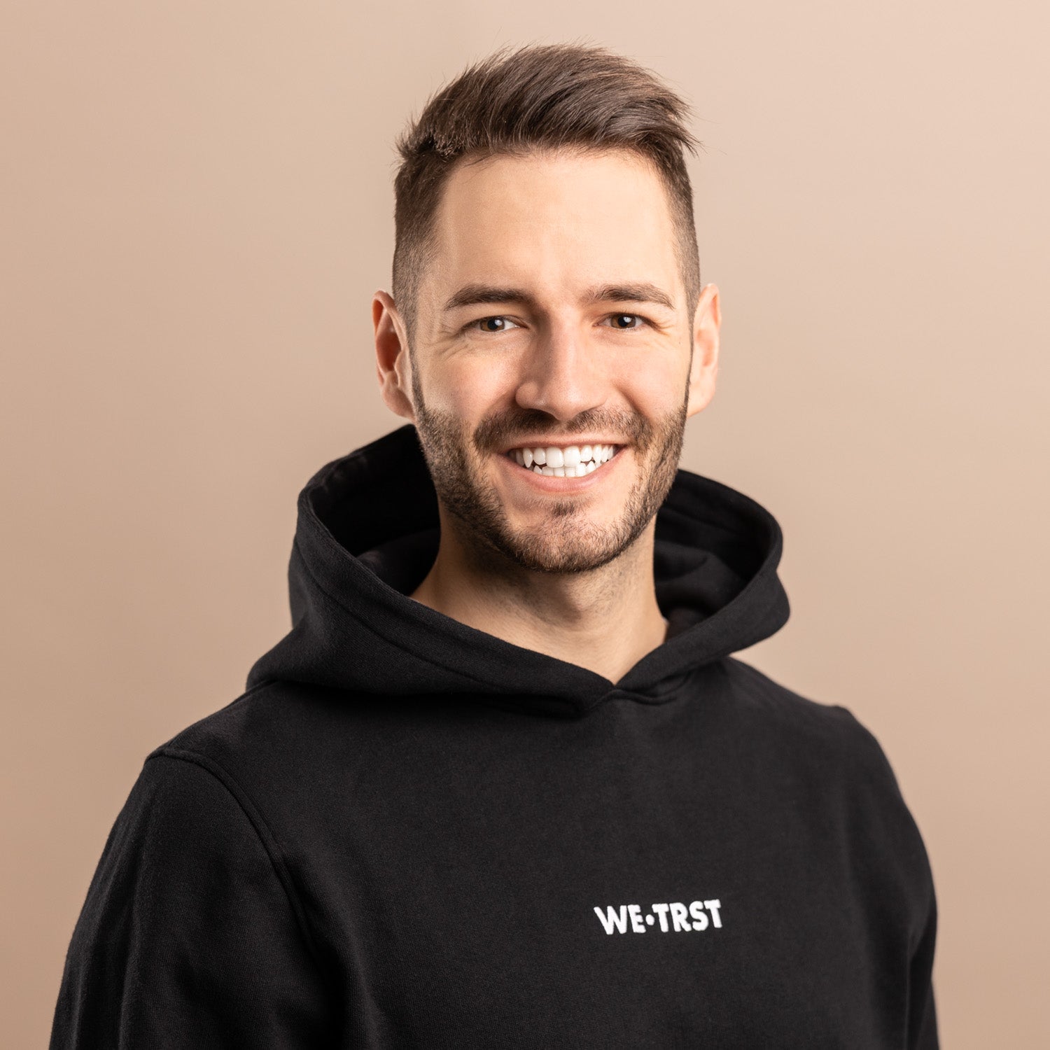 Das Bild zeigt Sandro Breu, den Mitgründer von WE•TRST. Er steht im Studio und blickt direkt in die Kamera. Er trägt einen schwarzen Hoodie der Marke, der mit dem markanten weißen Logo auf der Brust verziert ist.