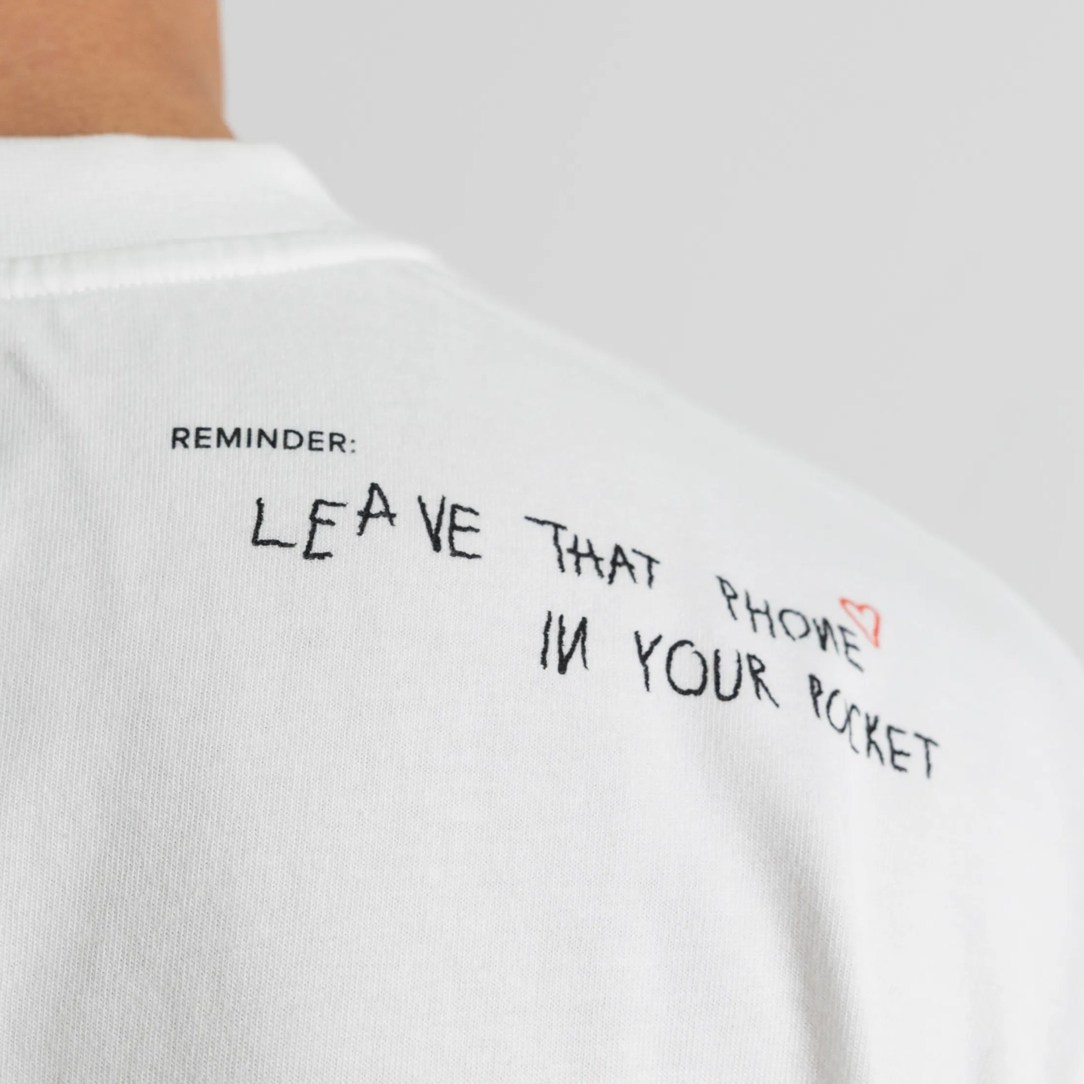 Detailaufnahme des Sticktexts «Reminder: Leave that phone in your pocket» auf dem Schulterbereich eines weissen WE•TRST x Dangel Twins T-Shirts.