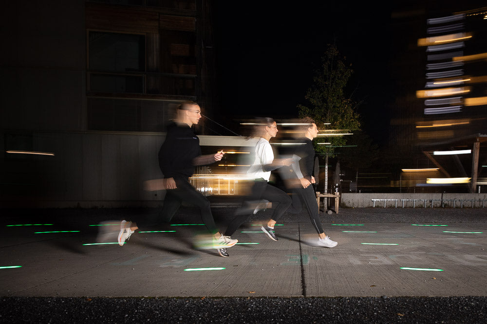 Three runners in motion at night, wearing WE•TRST athletic wear, captured with a motion blur effect in an urban environment with illuminated pathways.