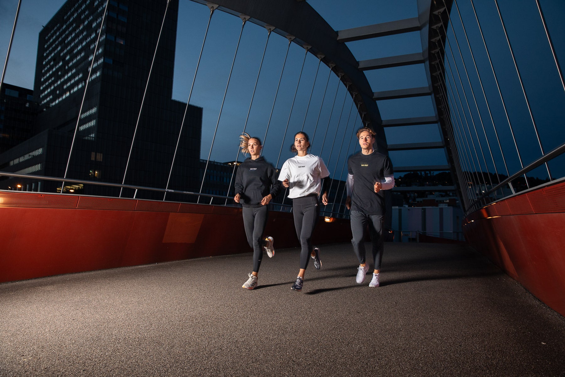 Drei Athleten, die nachts über eine moderne rote Brücke laufen und dabei stylische Activewear von WE•TRST Run Club tragen, mit beleuchteten Stadtgebäuden im Hintergrund.