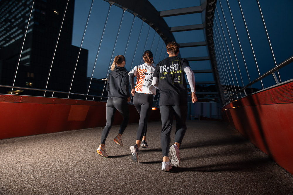 Drei Läufer in WE•TRST-Sportbekleidung joggen nachts über eine moderne Stadtbrücke, die von den Lichtern der Stadt beleuchtet wird. Die dynamische Bewegung und die stilvolle Sportkleidung unterstreichen einen aktiven Lebensstil, Nachtlauf und urbane Fitnesskultur.