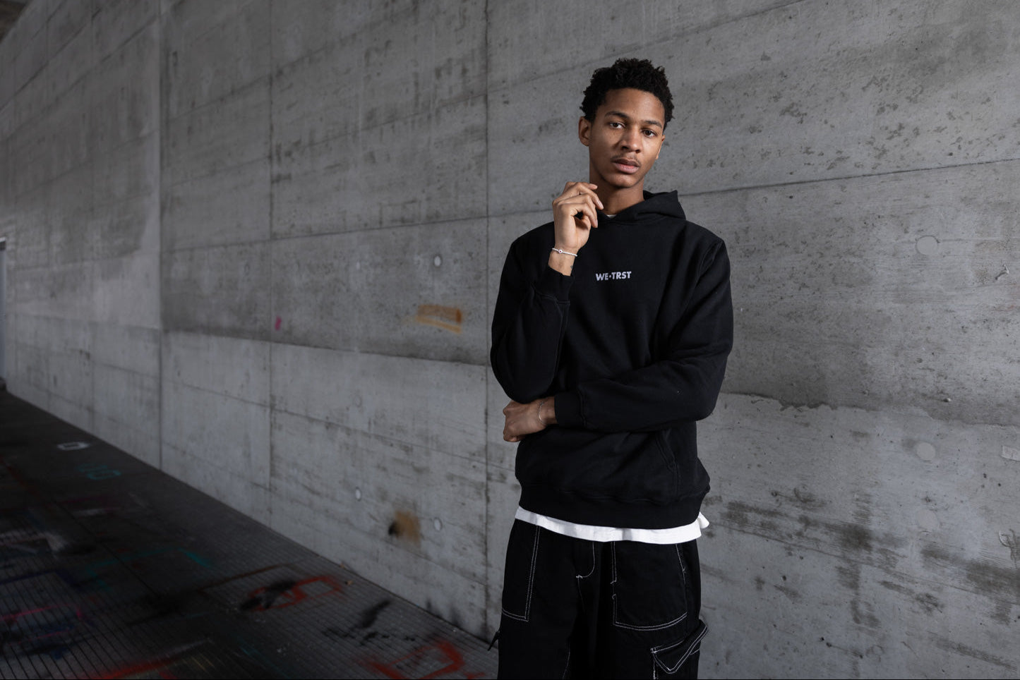 Männliches Model in schwarzem WE•TRST-Kapuzenpulli und Cargohose in einer urbanen Industriekulisse mit rohen Betonwänden, die für modernen, minimalistischen Streetwear-Stil und exklusive Mode steht.