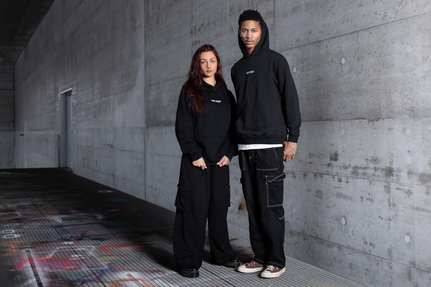Zwei Models in einer urbanen Industriekulisse mit passenden schwarzen WE•TRST Kapuzenpullis und übergroßen Cargohosen - moderner, minimalistischer Streetwear-Stil mit rohen Betonwänden und Graffiti-Elementen, der exklusive Mode widerspiegelt.