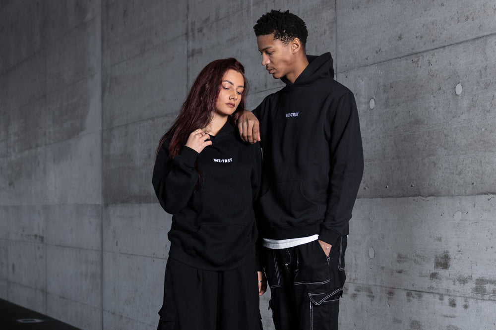 Zwei Models in einer urbanen Industriekulisse mit passenden schwarzen WE•TRST Kapuzenpullis und Cargohosen, die vor einer rohen Betonwand mit Graffiti-Elementen stehen und moderne, minimalistische Streetwear-Mode präsentieren.