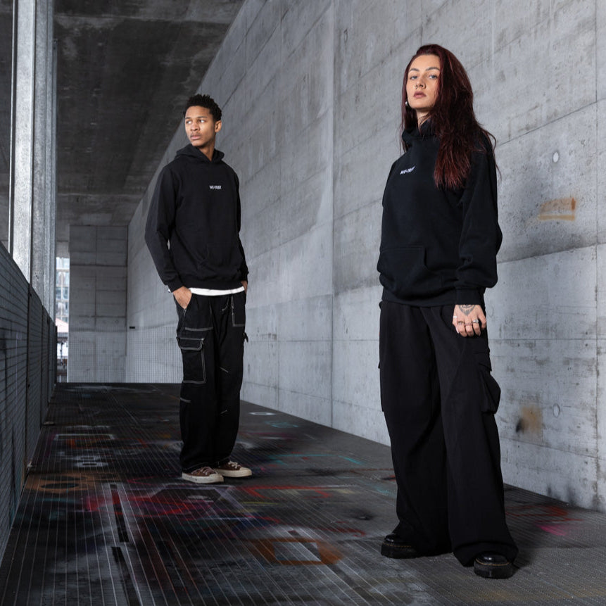 Two models in an industrial urban setting wearing black WE•TRST hoodies and oversized cargo pants – minimalist streetwear look with concrete walls and graffiti elements, representing exclusive, modern fashion.