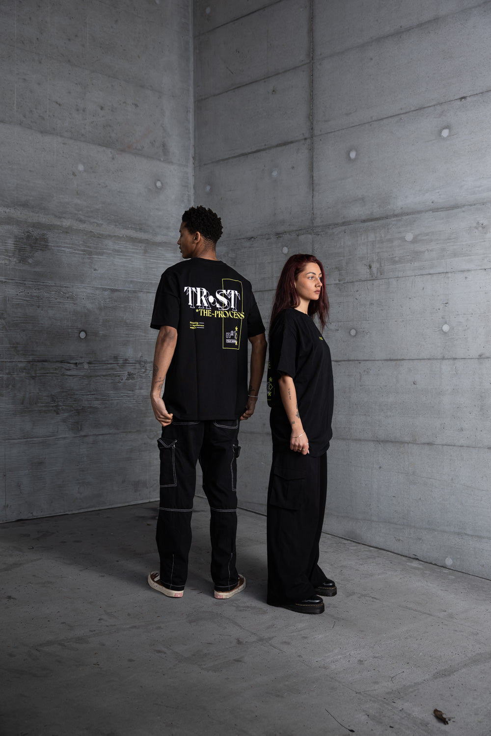 Two models in an urban setting wearing black WE•TRST oversized T-shirts with statement print on the back and minimalist cargo pants – modern streetwear look against a concrete wall, representing exclusive, progressive fashion.