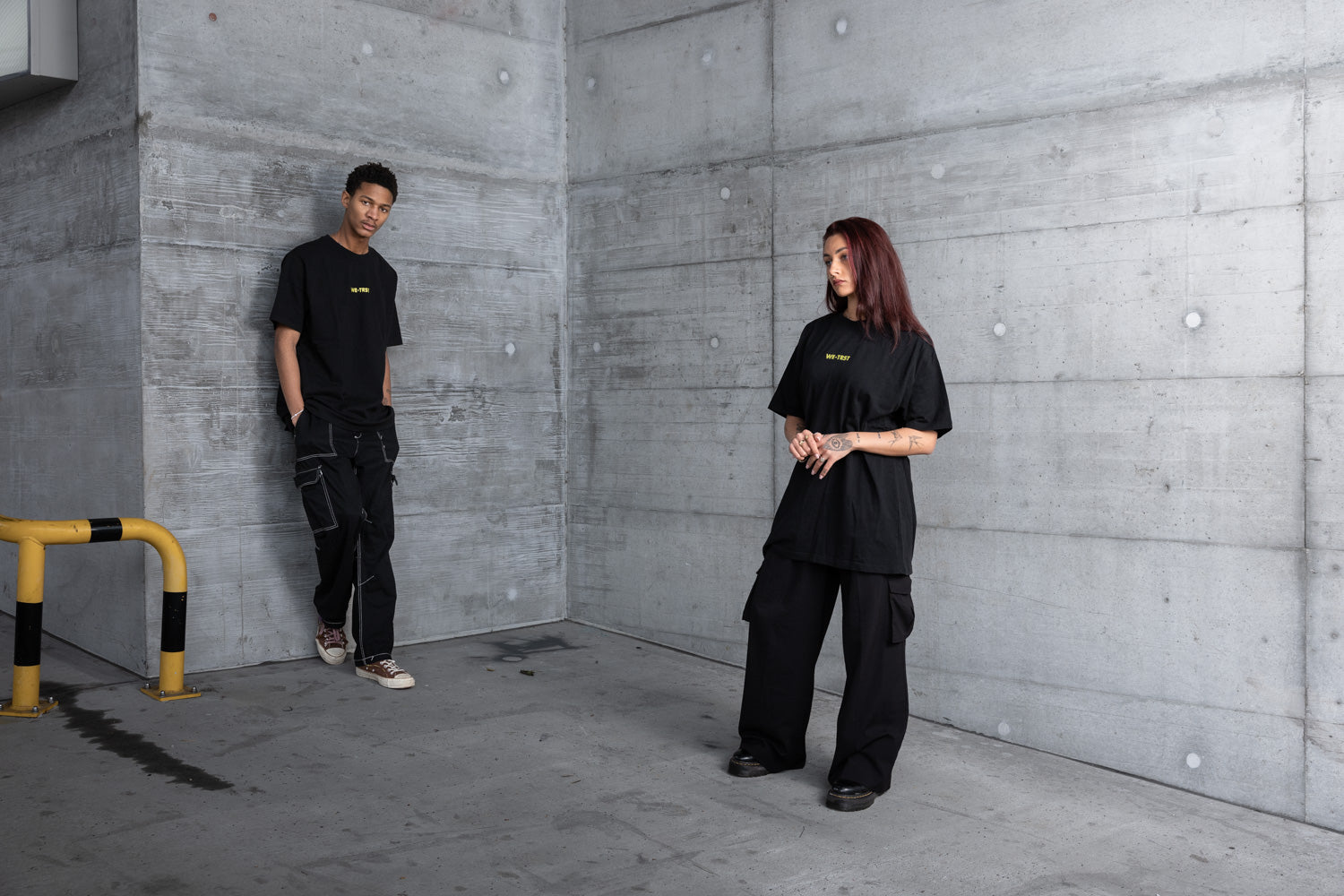 Zwei Models in einer urbanen Industriekulisse in schwarzen WE•TRST Oversized-T-Shirts und Cargohosen - minimalistischer Streetwear-Look vor rohen Betonwänden, der moderne, exklusive Mode widerspiegelt.