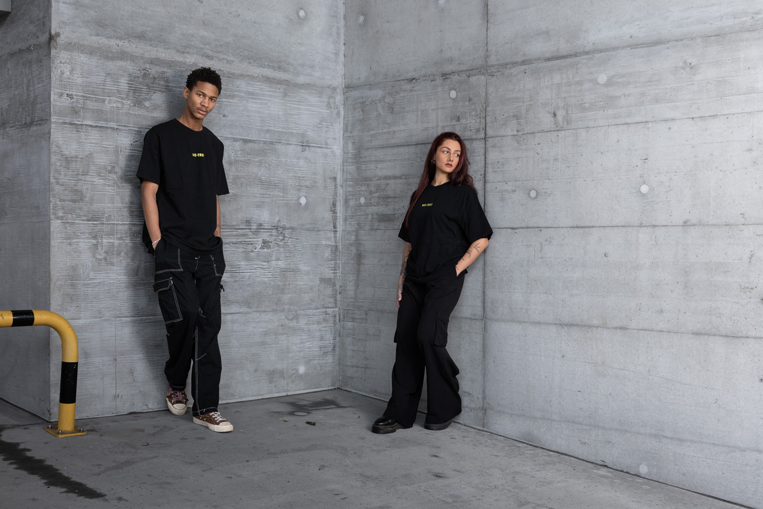 Zwei Models tragen schwarze WE•TRST Oversized-T-Shirts und Cargohosen in einer urbanen Industriekulisse mit rohen Betonwänden, die moderne, minimalistische Streetwear-Mode repräsentieren.