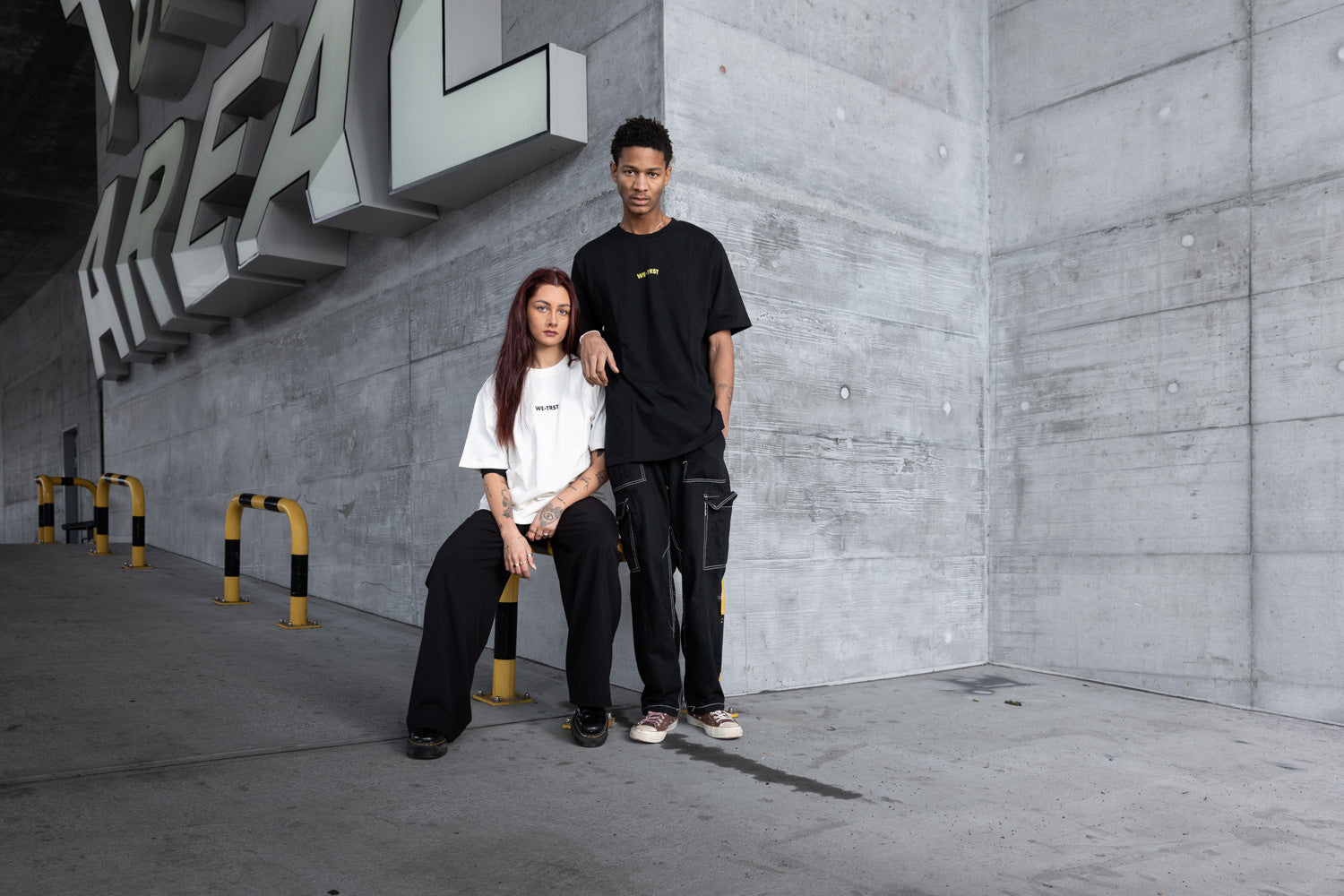 Zwei Models in einer urbanen Industriekulisse, das eine in einem weißen WE•TRST Oversized-T-Shirt, das andere in einem schwarzen WE•TRST T-Shirt mit Cargohose - moderner Streetwear-Stil vor einer rohen Betonwand mit kühnen architektonischen Elementen, die für exklusive Mode stehen.