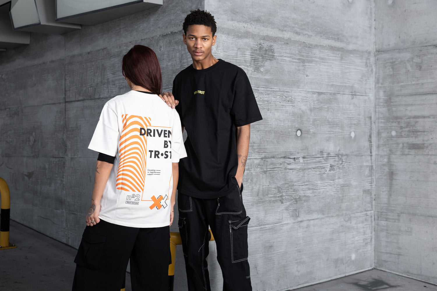 Zwei Models in einer urbanen Industriekulisse, die WE•TRST Streetwear tragen; das eine in einem weißen Oversized-T-Shirt mit auffälliger orangefarbener Grafik und Statement-Print, das andere in einem schwarzen Oversized-T-Shirt und einer Cargohose, die für moderne, exklusive Mode stehen.