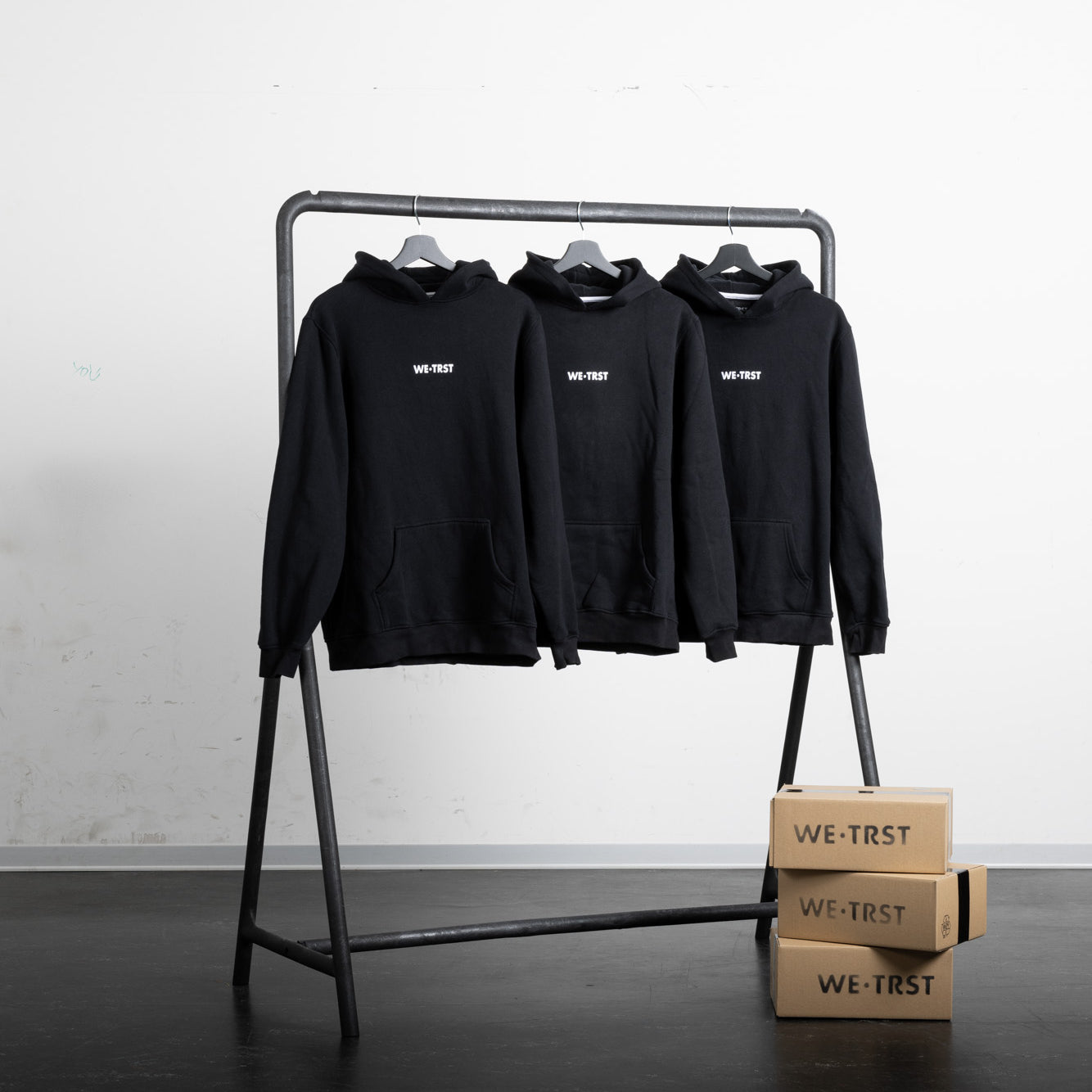 Drei schwarze WE•TRST-Kapuzenpullover hängen an einem minimalistischen industriellen Kleiderständer in einer sauberen, modernen Umgebung. Neben dem Regal stehen gestapelte Kartons mit dem WE•TRST-Logo, die das Branding und die Verpackung betonen.