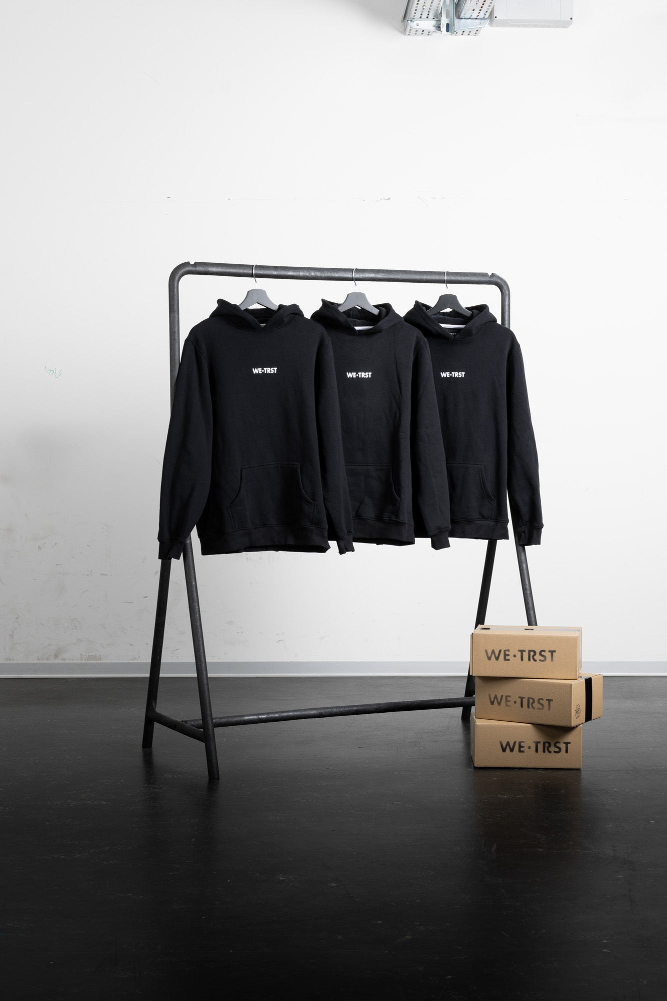 Three black WE•TRST hoodies hanging on a minimalist industrial clothing rack in a clean, modern setting. Stacked cardboard boxes with the WE•TRST logo are placed beside the rack, emphasizing branding and packaging.