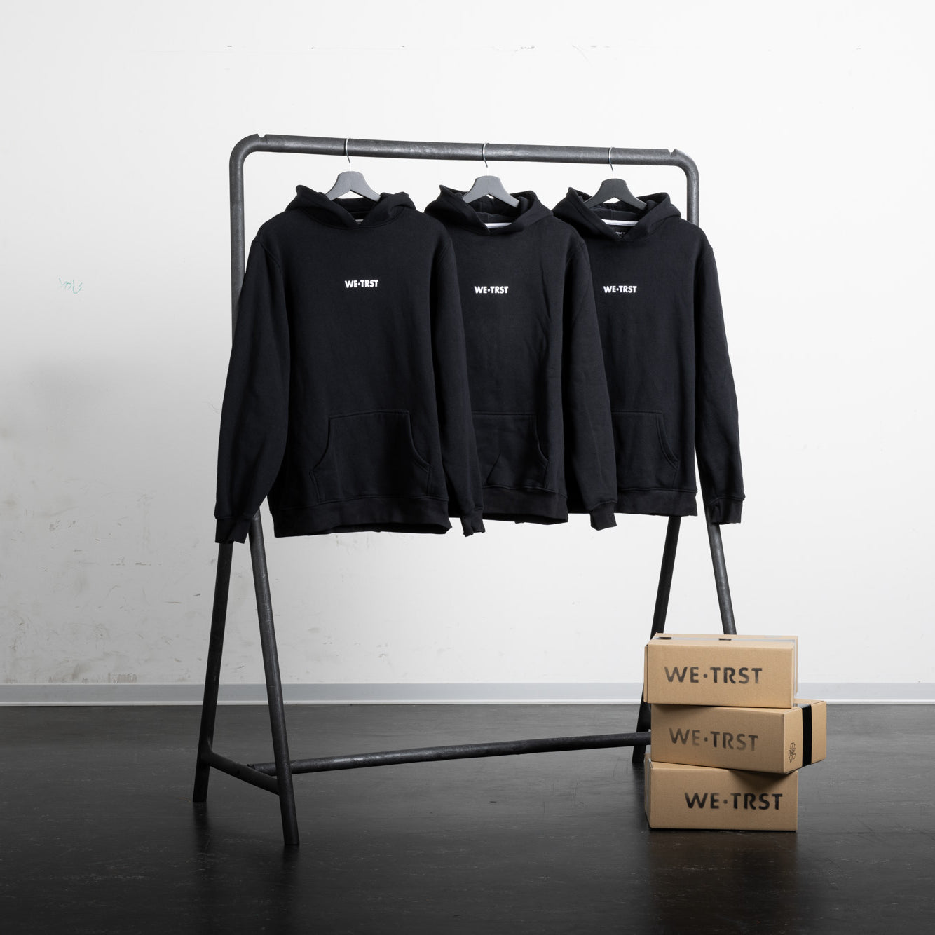 Drei schwarze Hoodies von WE•TRST an einem Kleiderständer. Darunter befinden sich drei braune Verpackungsschachteln.