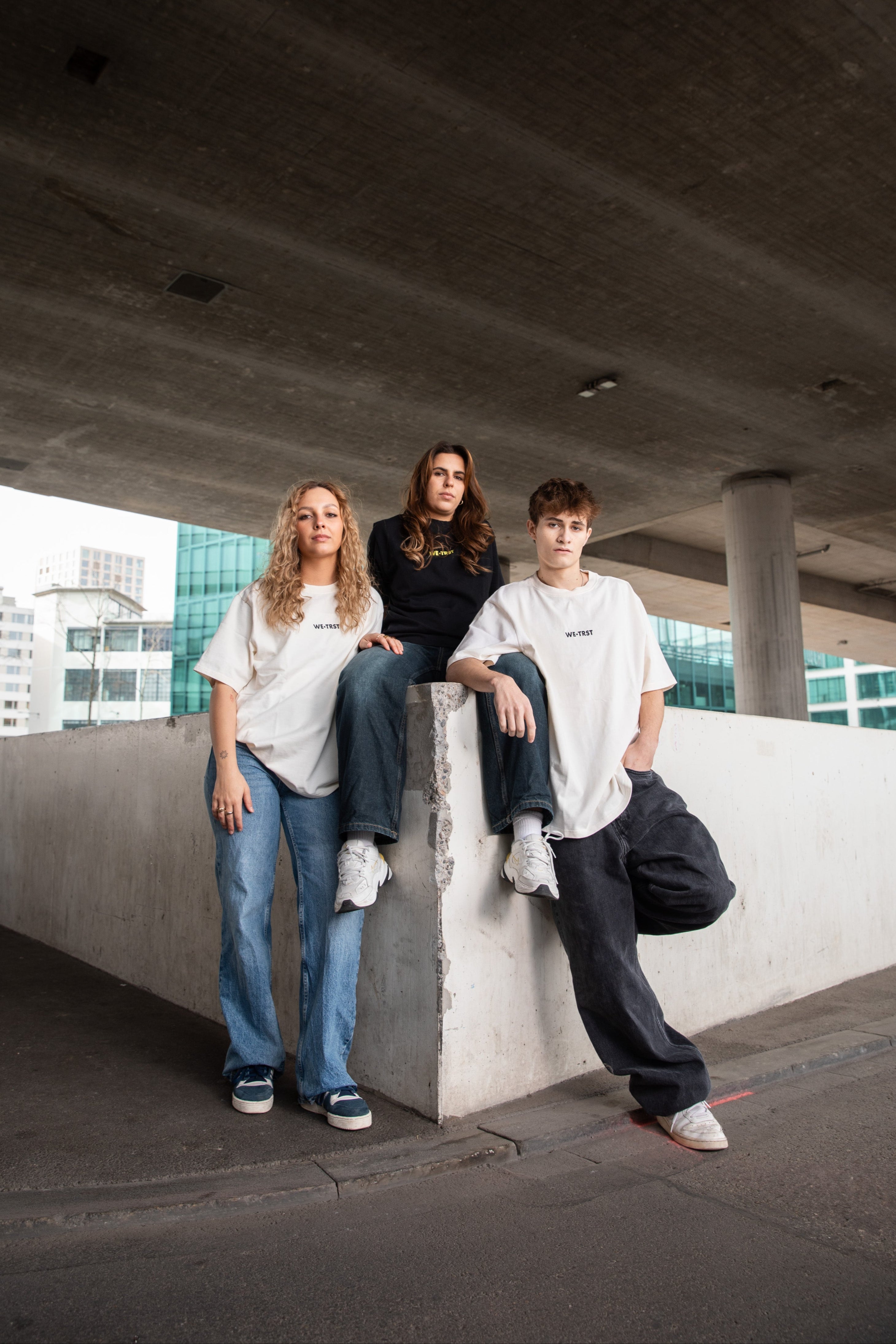 Three models in WE•TRST streetwear under a concrete bridge – urban fashion editorial with a minimalist aesthetic.