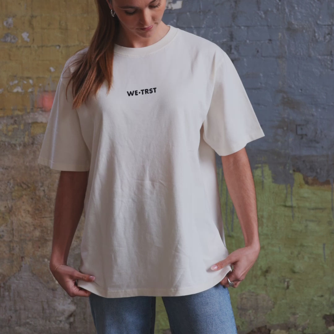 Ein weibliches Model trägt das WE•TRST T-Shirt „Driven by Trust“ in Off-White, das mit Jeans für einen lässigen Streetwear-Look gestylt wird.