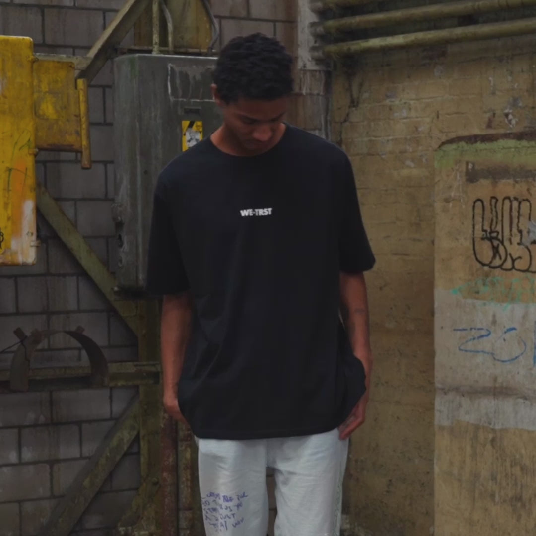 Männliches Model, das das WE•TRST Special Edition T-Shirt trägt, das sich durch einen cleanen Front-Look vor einem urbanen Industriehintergrund auszeichnet.