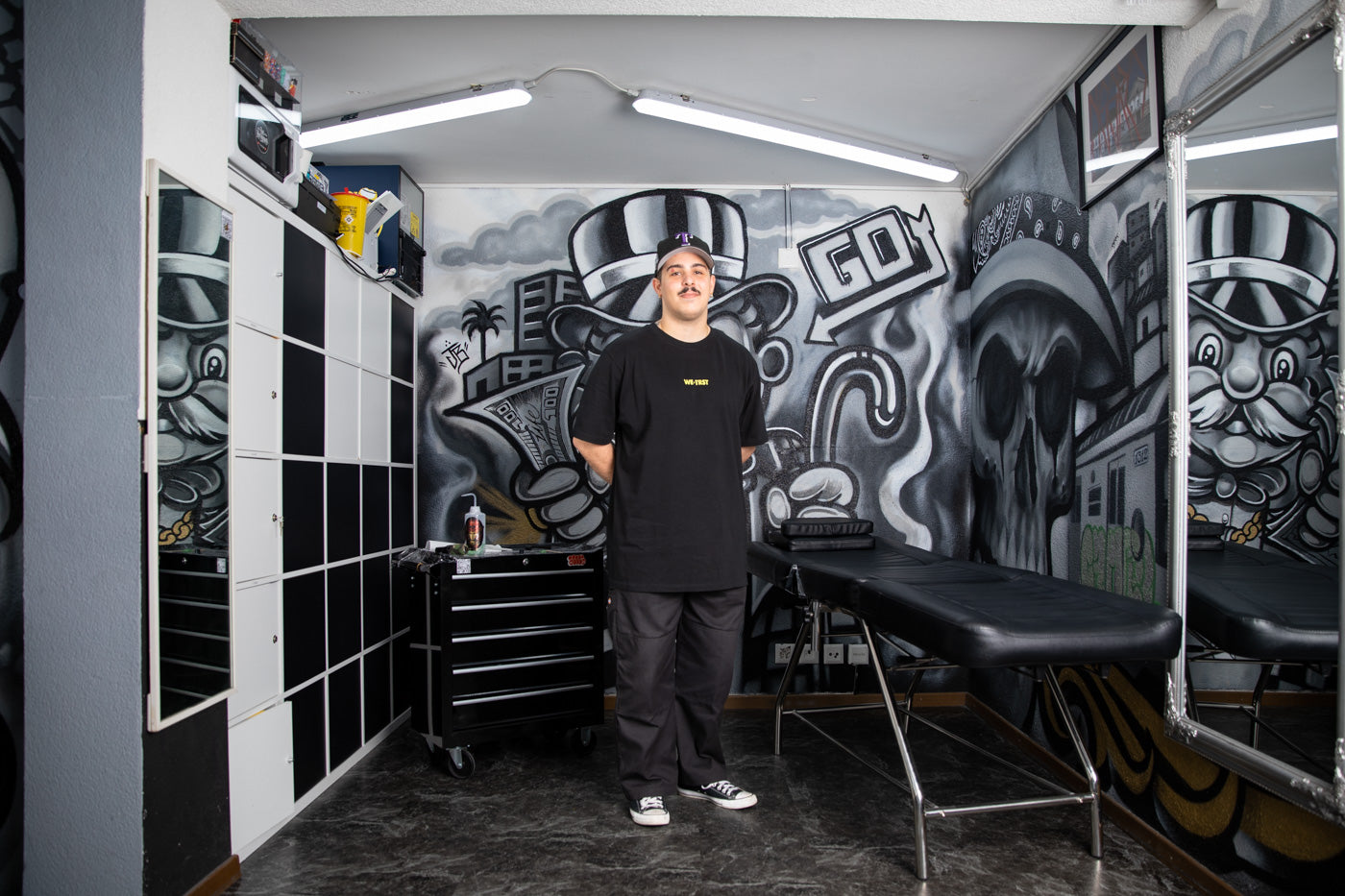 Ein junger Mann mit WE•TRST-T-Shirt steht stolz in seinem Tattoo-Studio.