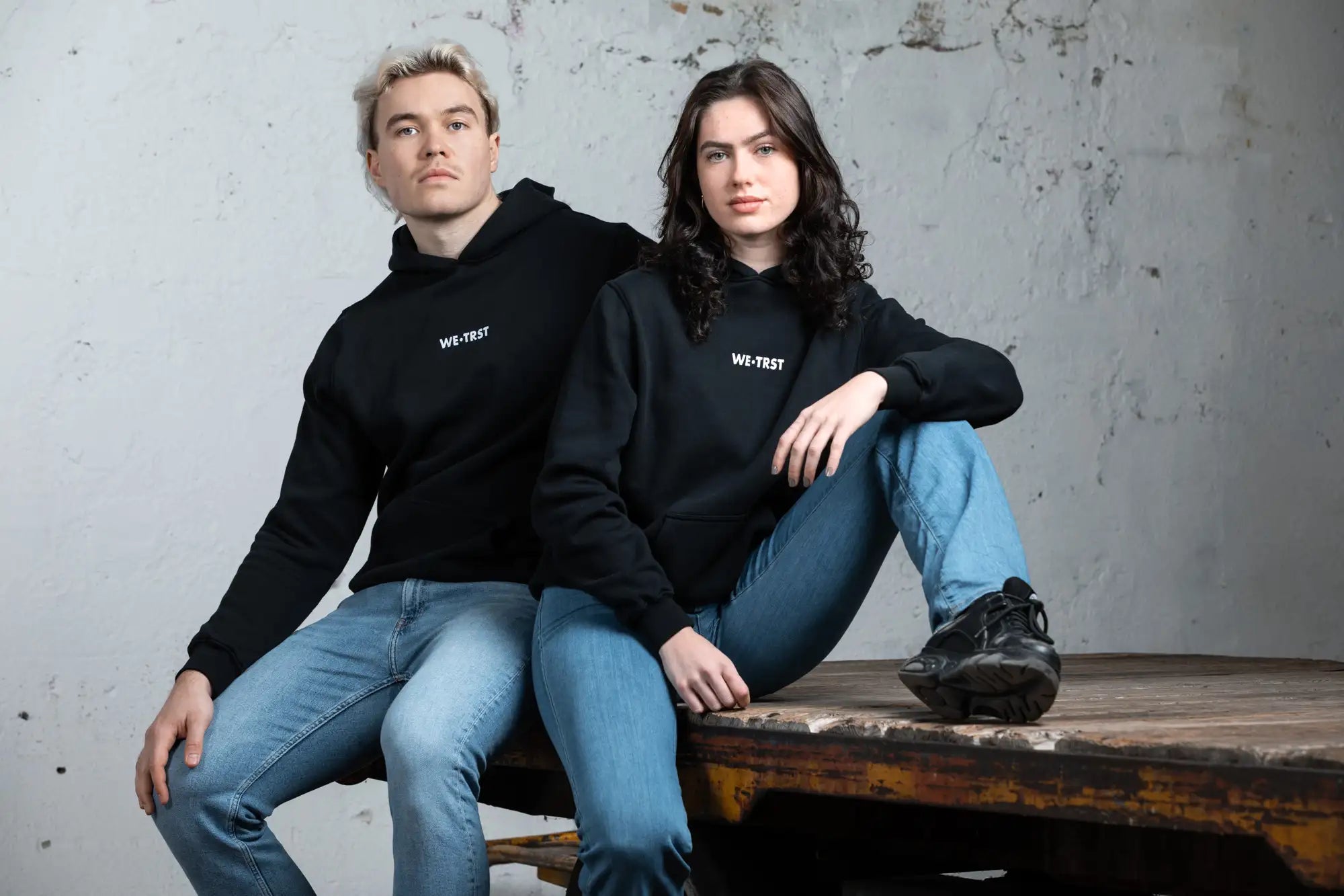 Ein männliches und ein weibliches Model in schwarzen WE•TRST-Kapuzenpullis sitzen lässig auf einem rustikalen Holztisch in einer industriellen Umgebung. Die Models tragen hellblaue Jeans und schwarze Turnschuhe, mit einem minimalistischen weißen Logo auf ihren Kapuzenpullovern.