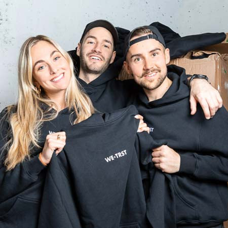 Das Bild zeigt die drei Gründer*innen von WE•TRST, die nebeneinander stehen und jeweils einen Hoodie der Marke tragen. Die drei Gründer*innen halten einen weiteren Hoodie hoch, um ihn zu präsentieren. Die Gründer*innen sehen stolz und zufrieden aus, während sie in die Kamera lächeln.