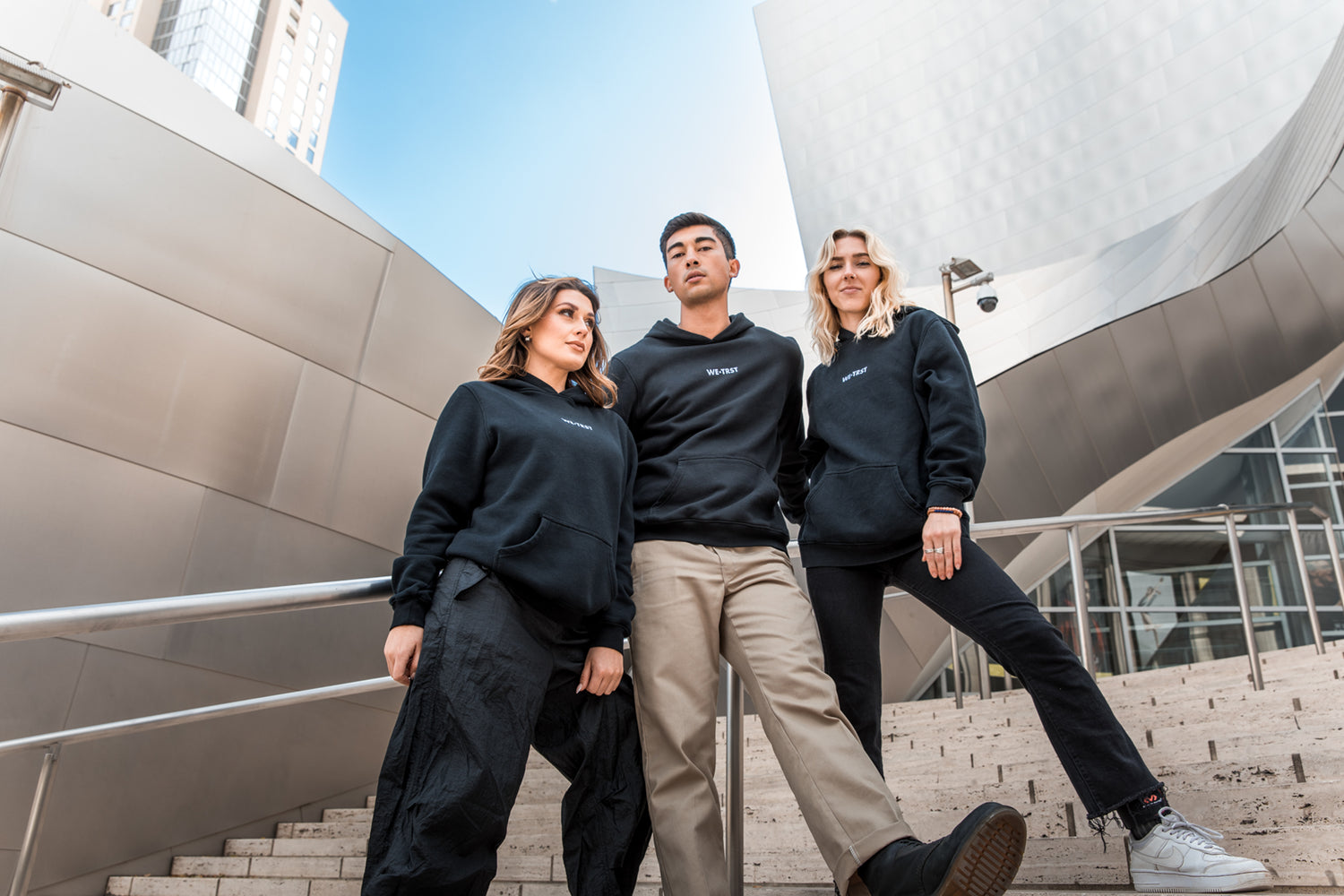 Drei Models stehen draußen vor einem Gebäude und tragen schwarze Hoodies von WE•TRST. Sie wirken selbstbewusst und stylish in ihren Outfits.