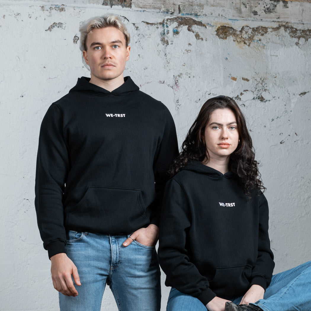Zwei Models, ein Mann und eine Frau, tragen schwarze WE•TRST-Kapuzenpullis in einer minimalistischen Industrieumgebung. Der Mann steht, während die Frau auf einem rustikalen Holztisch sitzt.