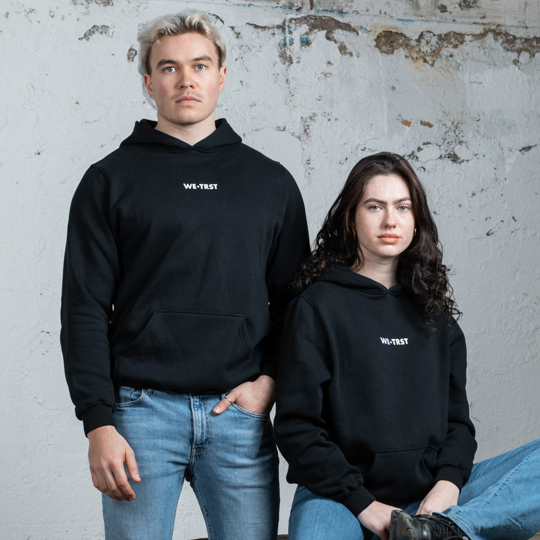 Zwei Models, ein Mann und eine Frau, tragen schwarze WE•TRST-Kapuzenpullis in einer minimalistischen Industrieumgebung. Der Mann steht, während die Frau auf einem rustikalen Holztisch sitzt.