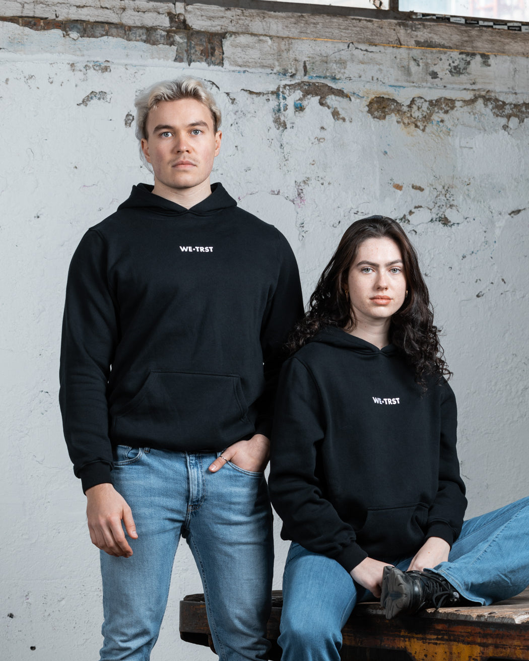 Zwei Models, ein Mann und eine Frau, tragen schwarze WE•TRST-Kapuzenpullis in einer minimalistischen Industrieumgebung. Der Mann steht, während die Frau auf einem rustikalen Holztisch sitzt.