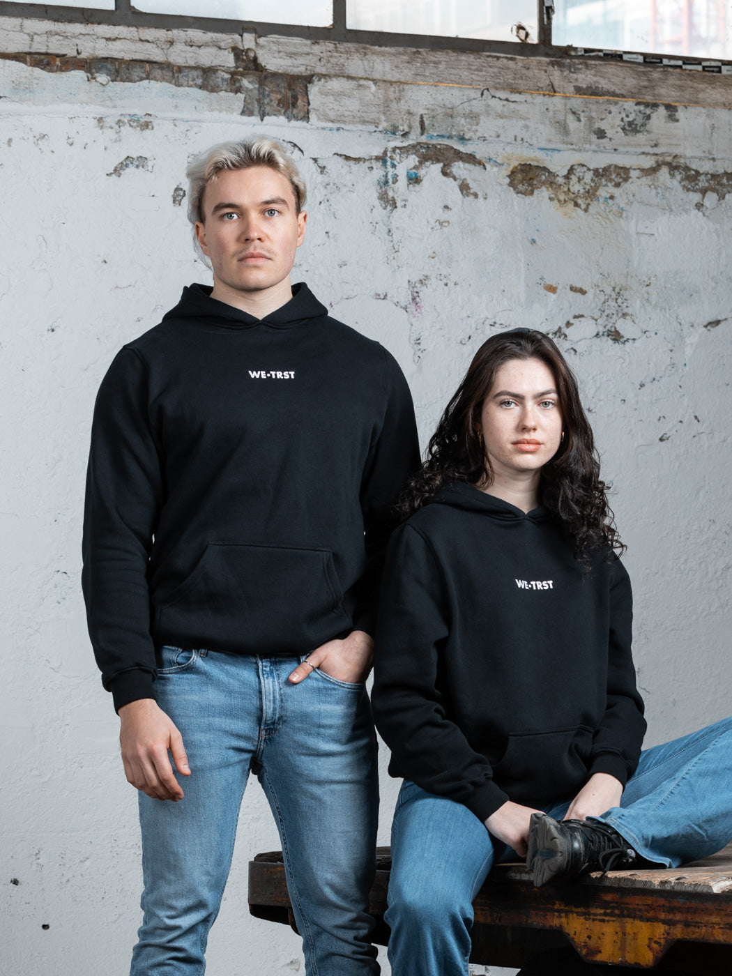 Zwei Models, ein Mann und eine Frau, tragen schwarze WE•TRST-Kapuzenpullis in einer minimalistischen Industrieumgebung. Der Mann steht, während die Frau auf einem rustikalen Holztisch sitzt.