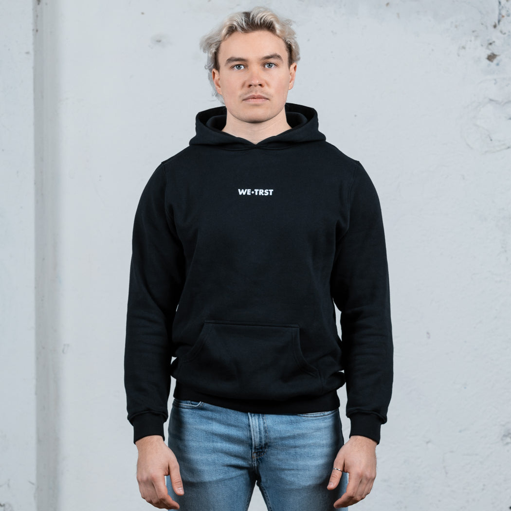 Ein männliches Modell mit platinblondem Haar, das einen schwarzen WE•TRST-Kapuzenpullover und hellblaue Jeans trägt und vor einer strukturierten weißen Wand steht.