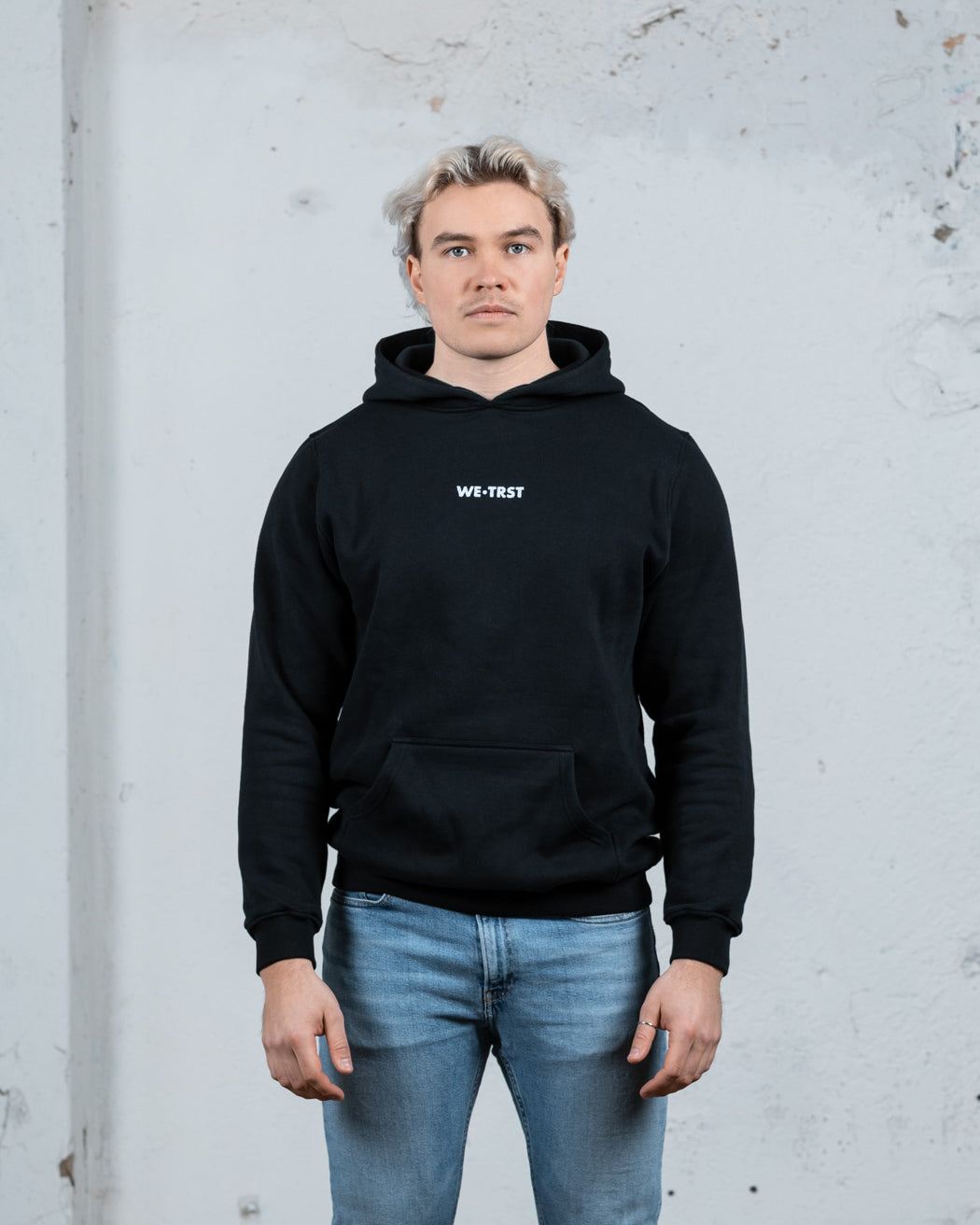 Ein männliches Modell mit platinblondem Haar, das einen schwarzen WE•TRST-Kapuzenpullover und hellblaue Jeans trägt und vor einer strukturierten weißen Wand steht.