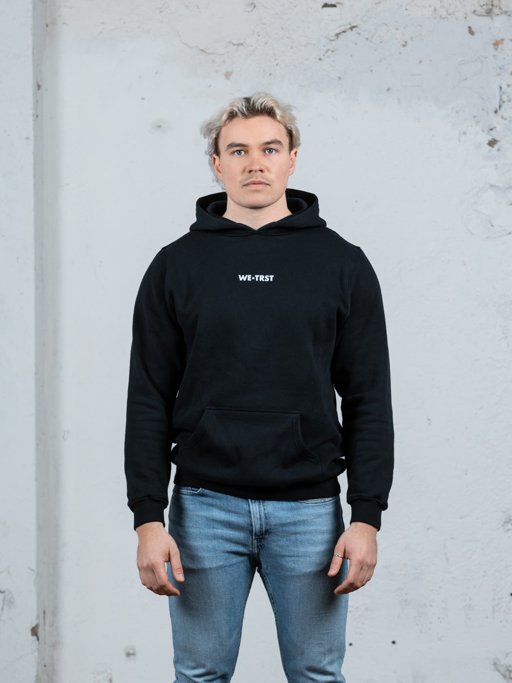 Ein männliches Modell mit platinblondem Haar, das einen schwarzen WE•TRST-Kapuzenpullover und hellblaue Jeans trägt und vor einer strukturierten weißen Wand steht.