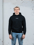 Ein männliches Modell mit platinblondem Haar, das einen schwarzen WE•TRST-Kapuzenpullover und hellblaue Jeans trägt und vor einer strukturierten weißen Wand steht.