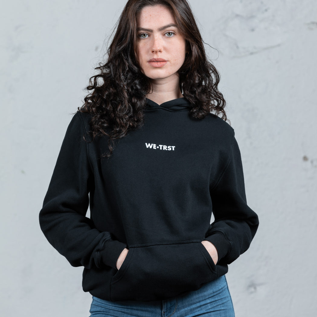 Ein weibliches Modell mit dunklem, lockigem Haar in einem schwarzen WE•TRST-Kapuzenpulli und blauen Jeans steht vor einer rohen, weißen Industriewand.