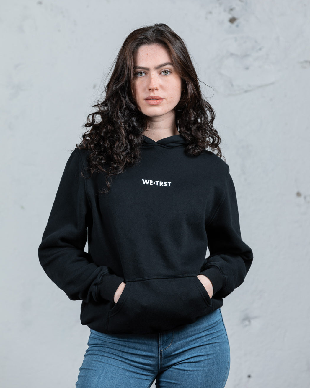 Ein weibliches Modell mit dunklem, lockigem Haar in einem schwarzen WE•TRST-Kapuzenpulli und blauen Jeans steht vor einer rohen, weißen Industriewand.