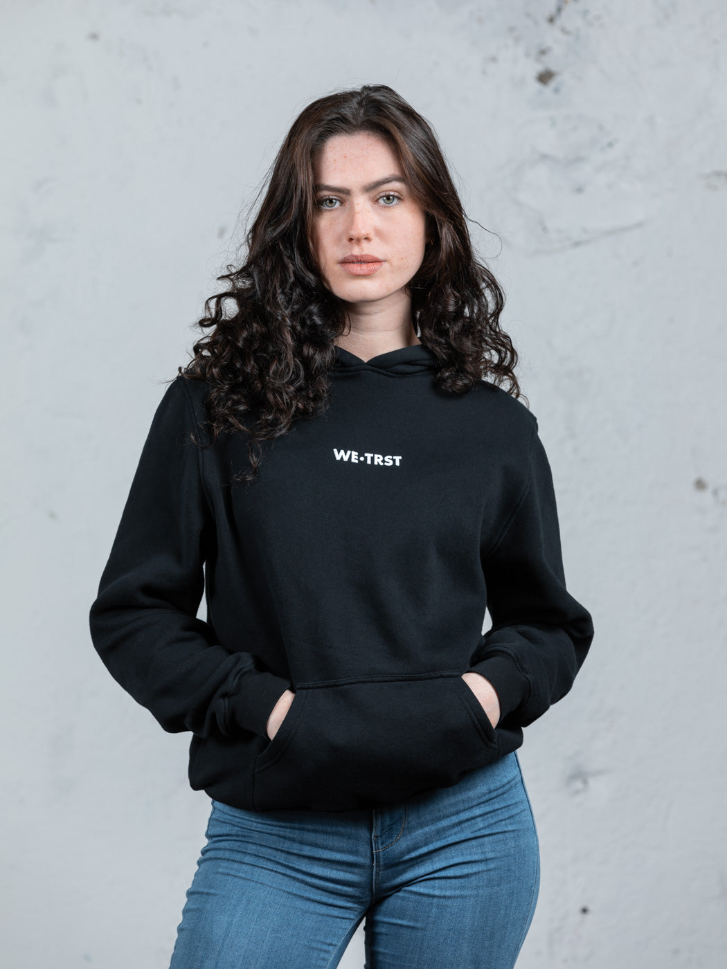 Ein weibliches Modell mit dunklem, lockigem Haar in einem schwarzen WE•TRST-Kapuzenpulli und blauen Jeans steht vor einer rohen, weißen Industriewand.