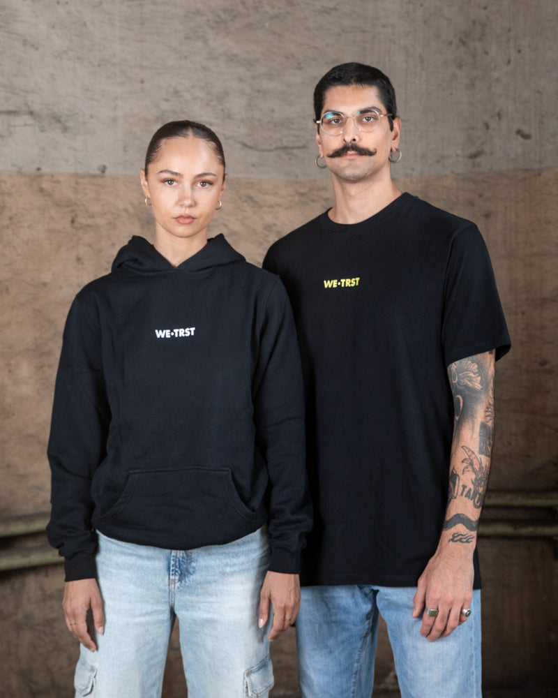 Zwei Models, eine Frau und ein Mann, tragen schwarze WE•TRST Streetwear in einer industriellen Umgebung. Die Frau trägt einen Kapuzenpulli, der Mann ein T-Shirt, beide mit minimalistischen Logos versehen.