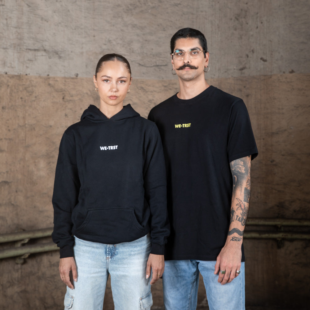 Zwei Models, eine Frau und ein Mann, tragen schwarze WE•TRST Streetwear in einer industriellen Umgebung. Die Frau trägt einen Kapuzenpulli, der Mann ein T-Shirt, beide mit minimalistischen Logos versehen.