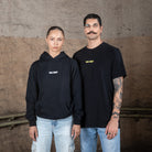 Zwei Models, eine Frau und ein Mann, tragen schwarze WE•TRST Streetwear in einer industriellen Umgebung. Die Frau trägt einen Kapuzenpulli, der Mann ein T-Shirt, beide mit minimalistischen Logos versehen.
