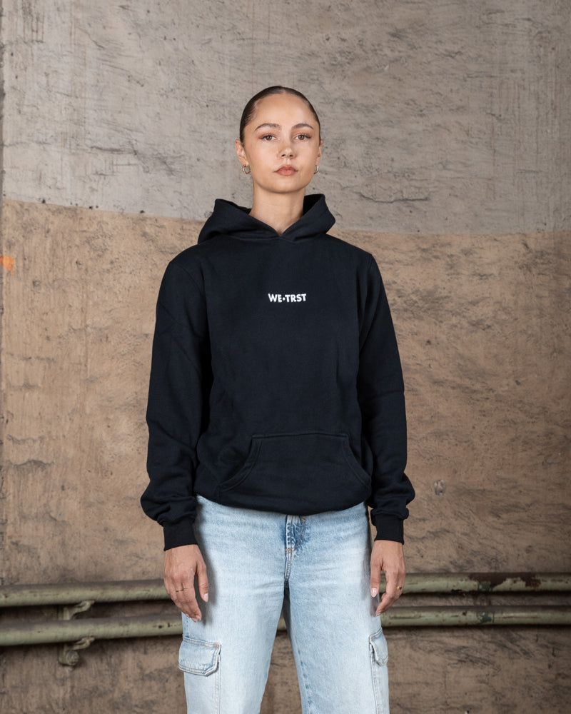 Ein weibliches Model in einem schwarzen WE•TRST-Kapuzenpulli mit weißem Branding auf der Brust, gestylt mit hellen Cargo-Jeans, steht vor einem rauen Betonhintergrund.